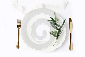 Festive table summer setting with golden cutlery, olive branch, porcelain dinner plate and silk ribbon on white table