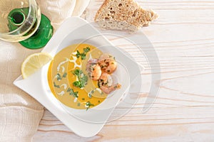 Festive vegetable cream soup from red kuri squash with shrimps and parsley garnish in an elegant white plate, bread and wine on a