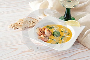 Festive vegetable cream soup from red kuri squash with shrimps and parsley garnish in a white plate, bread and wine on a light