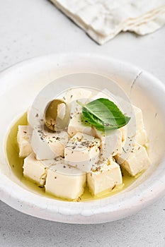 Feta cheese cubes with rosemary, olives and olive oil sauce in white bowl on light gray background. Traditional Greek homemade