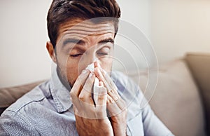 This fever just wont go away. a handsome young man blowing his nose with a tissue while relaxing on a sofa at home.