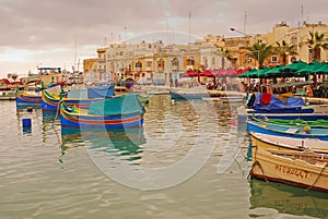 Few colourful traditional Maltese fishing boats Luzzu docking with old houses behind at cloudy fishing village Marsaxlokk, Malta