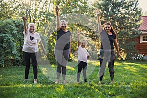 Few generations of family smiling females standing on green grass holding one hand up stretching warming up on meadow