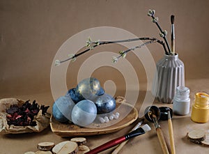 Few painted chicken eggs on wooden plate, dried red petals on crumpled paper and tree branch and paint brush in little ceramic