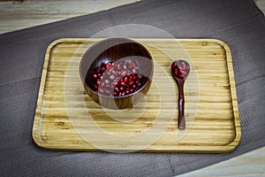 A few pomegranate seeds in a wooden bowl. Next to the wooden bowl is a wooden spoon with a few seeds in it. Wooden tray. The