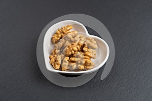 A few shelled walnuts lie in a saucer in the shape of a heart on textured slate board