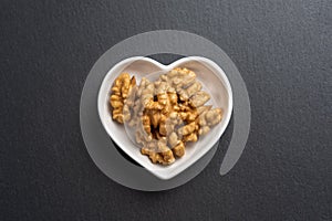 A few shelled walnuts lie in a saucer in the shape of a heart on textured slate board