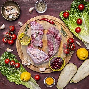 Few slices of raw beef on a cutting board, around lie ingredients, fresh vegetables wooden rustic background top view