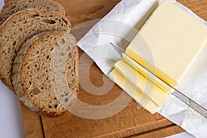 A few slices of yellow butter cut off from a large piece with a knife on a brown wooden cutting board. Several slices of rye bread