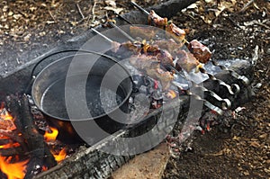 Field kitchen tourist camp on open fire of the bonfire, boiling water in boiler and grilled pork meat skewers