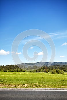 Field by a road with blue sky and the mountain forest in the dis