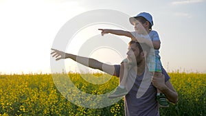 Field walk of dad and son in sunny weather, men with little boy go by field and point hand to horizon,