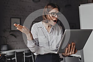 Fierce business lady looks indignantly and shocked into laptop and spreads her arms against background of white