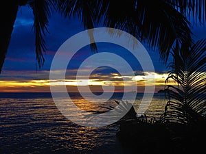 Fiery glow sunset over a beautiful tropical beach and ocean water. Different colours of clouds and the silhouette of palm tree.