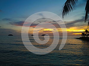 Fiery glow sunset over a beautiful tropical beach and ocean water. Different colours of clouds and the silhouette of palm tree.