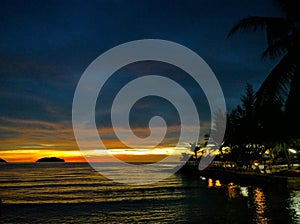 Fiery glow sunset over a beautiful tropical beach and ocean water. Different colours of clouds and the silhouette of palm tree.