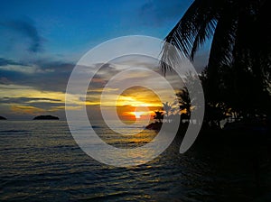 Fiery glow sunset over a beautiful tropical beach and ocean water. Different colours of clouds and the silhouette of palm tree.