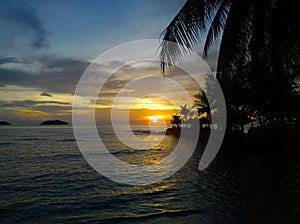 Fiery glow sunset over a beautiful tropical beach and ocean water. Different colours of clouds and the silhouette of palm tree.