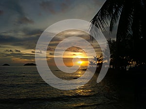 Fiery glow sunset over a beautiful tropical beach and ocean water. Different colours of clouds and the silhouette of palm tree.