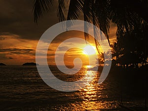 Fiery glow sunset over a beautiful tropical beach and ocean water. Different colours of clouds and the silhouette of palm tree.