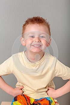 Fiery redhead young boy in yellow shirt fists on hips sitting on a stool with a huge cheeky grin. Portrait on grey background.