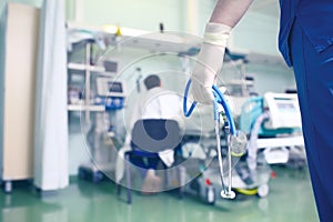 Figure of a doctor with stethoscope in hands on the background of a man siiting near the patient bed in hospital ward