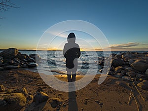 Figure of girl in warm hooded winter jacket on lake at sunset in late autumn, a loving landscape in nature