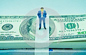 Figure of a man in a suit on top of a wad of hundred dollar bills in a white background