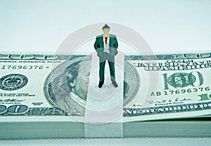 Figure of a man in a suit on top of a wad of hundred dollar bills in a white background