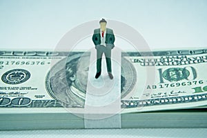 Figure of a man in a suit on top of a wad of hundred dollar bills in a white background