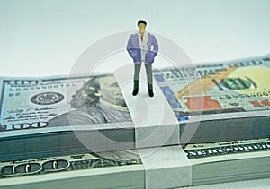 Figure of a man in a suit on top of a wad of hundred dollar bills in a white background