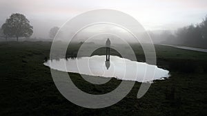 A figure standing reflected in a pond, silhouetted on a spooky foggy winters evening in the countryside