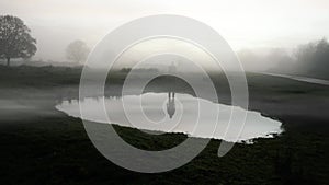 A figure standing reflected in a pond, silhouetted on a spooky foggy winters evening in the countryside