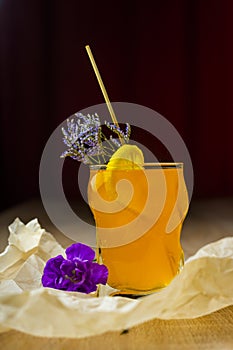 Figured glass of yellow fruit juice with a slice of lemon, a green branch of small flowers and a purple flower beside, on crumple