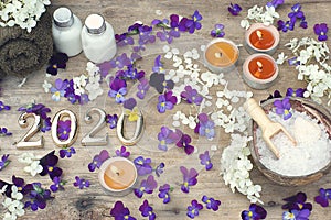 Figures 2020 on a wooden background next to purple pansy flowers, burning candles and white bath salt, spa concept