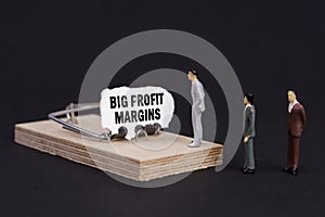Figures of businessmen stand near a mousetrap, the bait in which is torn paper with the inscription - Big Profit Margins