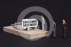 Figures of businessmen stand near a mousetrap, the bait in which is torn paper with the inscription - Consumer Sentiment