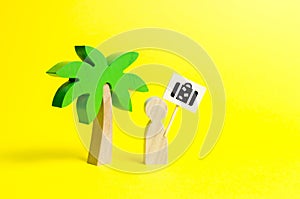 Figurine of a man with a poster with a bag under a palm tree on a yellow background. Concept of wanting a vacation trip photo