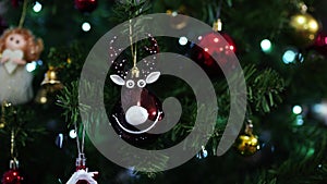Figurine of a smiling deer hangs on a green branch of a Christmas tree among the balls