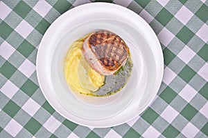 Filet mignon with mashed potatoes and pesto sauce served in a white plate over green plaid tablecloth. Beef steak.