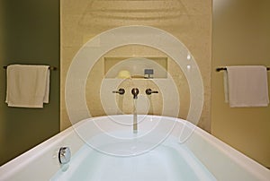 Filled white bathtub with brass tap mounted on the marble wall and shower screens on both sides