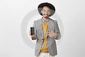 Finally new model of smartphone. Positive happy guy in stylish outfit and hat, showing smartphone and pointing at device