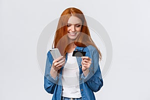 Finance, business and education concept. Cheerful cute alluring redhead woman making online purchase, holding smartphone