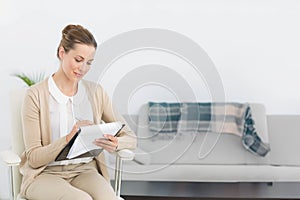 Female counselor wearing businesswear writing notes on clipboard sitting in office near sofa, plant