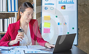 Financial, Asian businesswoman in red suit holding cup of coffee sitting on desk in office, having computer for doing accounting