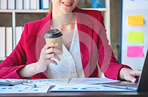 Financial, Asian businesswoman in red suit holding cup of coffee sitting on desk in office, having computer for doing accounting