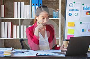 Financial, Asian businesswoman in red suit holding cup of coffee sitting on desk in office, having computer for doing accounting