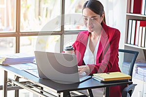 Financial, Asian businesswoman in red suit holding cup of coffee sitting on desk in office, having computer for doing accounting