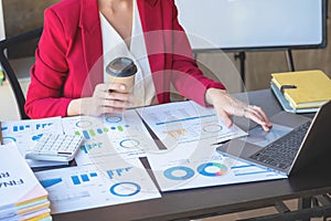 Financial, Asian businesswoman in red suit holding cup of coffee sitting on desk in office, having computer for doing accounting