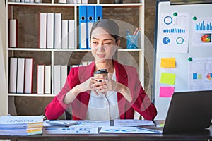 Financial, Asian businesswoman in red suit holding cup of coffee sitting on desk in office, having computer for doing accounting